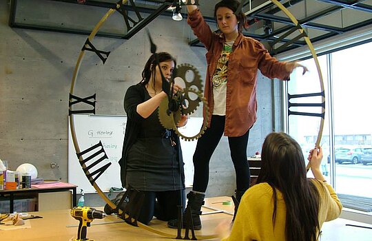 Des étudiantes debout sur une table en classe montent une horloge géante.