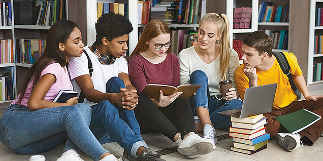 Cinq étudiants assis par terre dans une bibliothèque observent un livre.