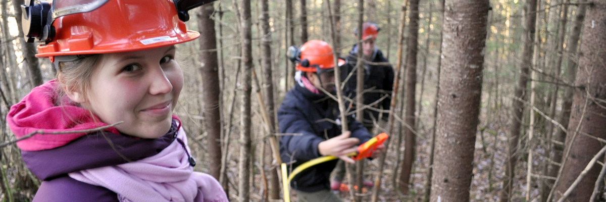 Une étudiante sourit, elle est en forêt avec un casque de protection sur la tête.