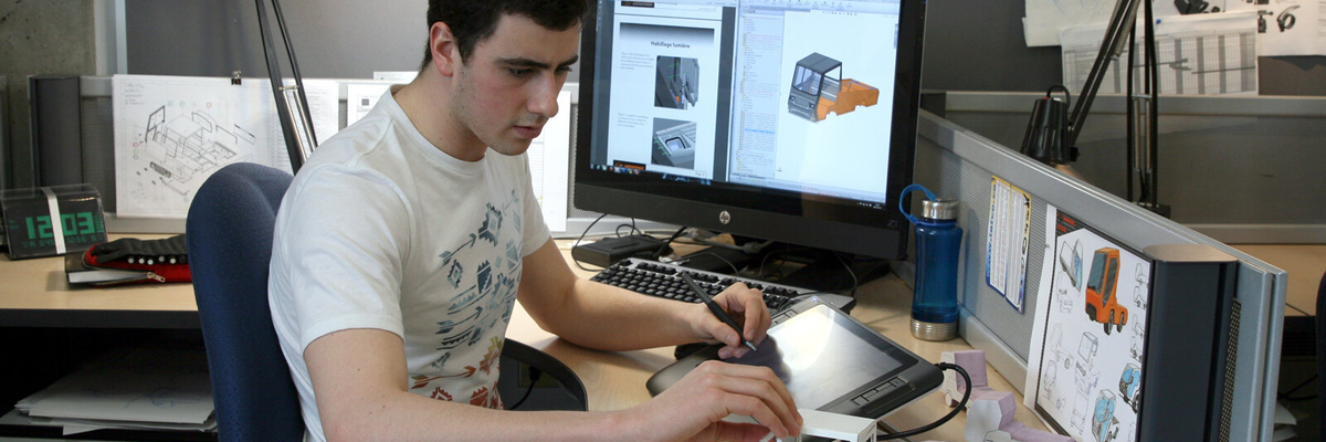 Un étudiant dessine sur une tablette électronique tout en observant des maquettes de papier installées devant lui.lés